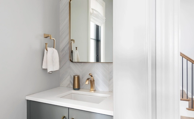 Modern bathroom vanity with gold faucet, white countertops, and herringbone tile backsplash above the vanity.